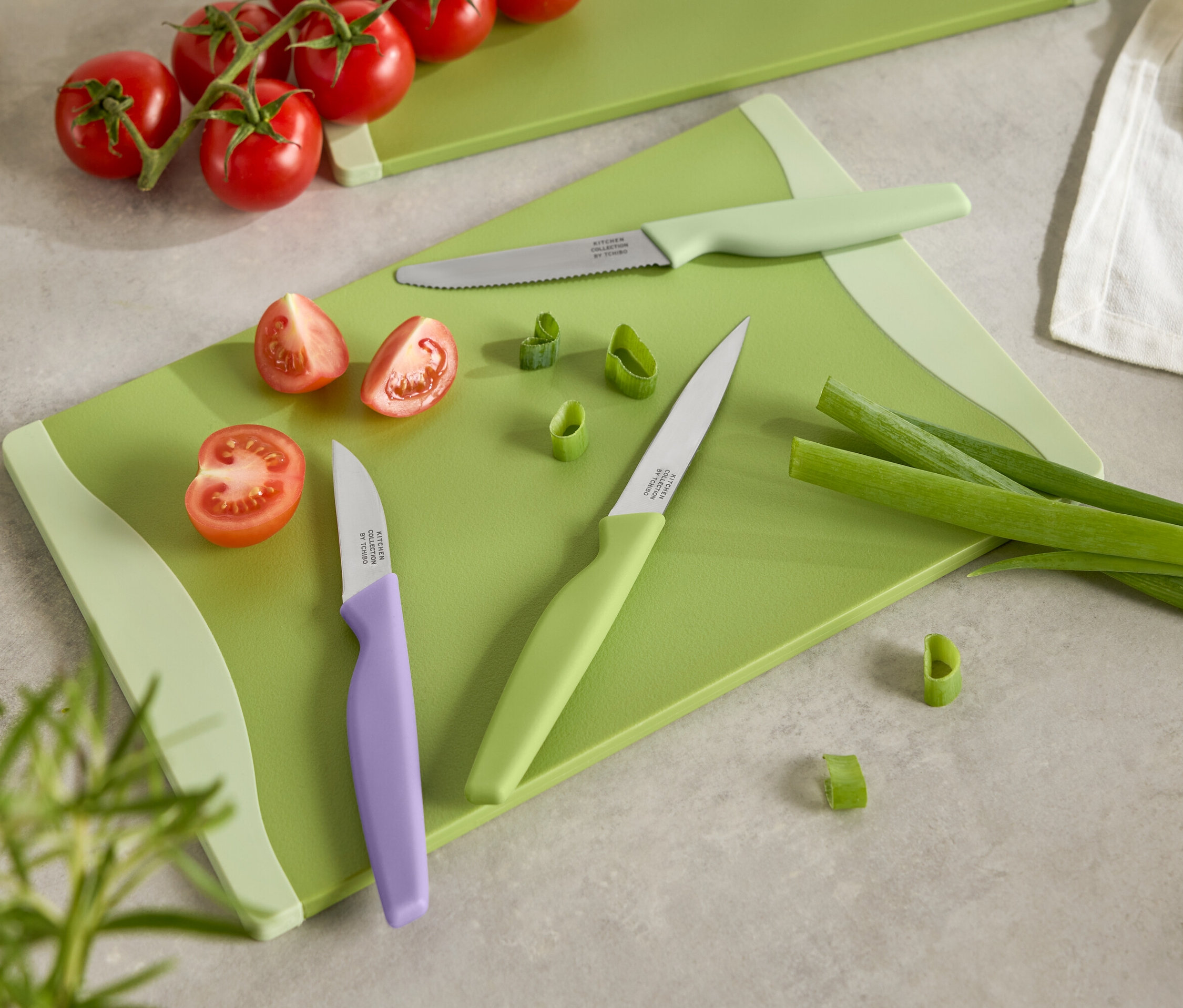 Zielona deska do krojenia z pokrojonymi pomidorami, dymką i trzema nożami z kolorowymi rączkami.