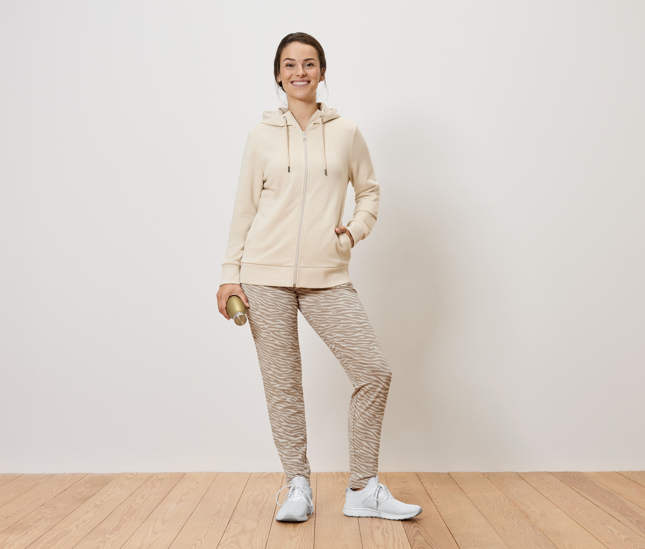 Kobieta stoi i trzyma złotą butelkę. Ma na sobie beżową bluzę z kapturem na zamek błyskawiczny, wzorzyste spodnie i białe trampki.