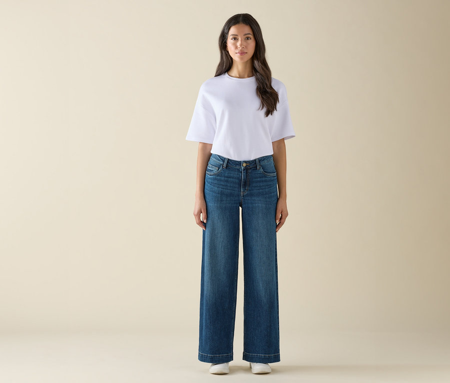 Kobieta stoi w białym t-shircie, szerokich niebieskich dżinsach i białych tenisówkach.