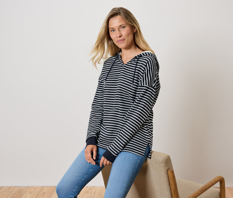 Kobieta siedzi na krześle, ubrana w niebieskie dżinsy i bluzę w paski z kapturem.