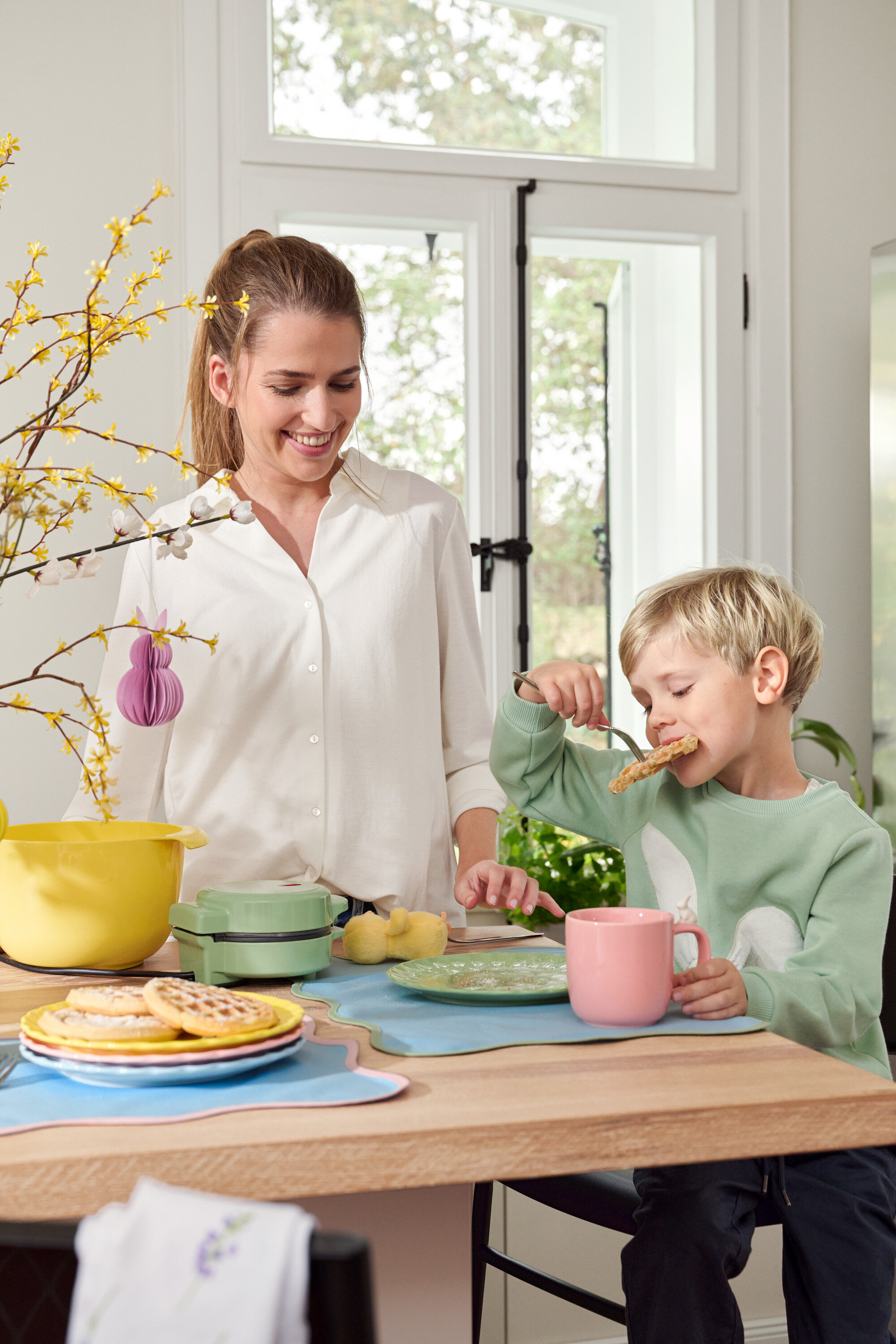 Kobieta i dziecko przy stole. Dziecko je gofra widelcem, kobieta się uśmiecha.