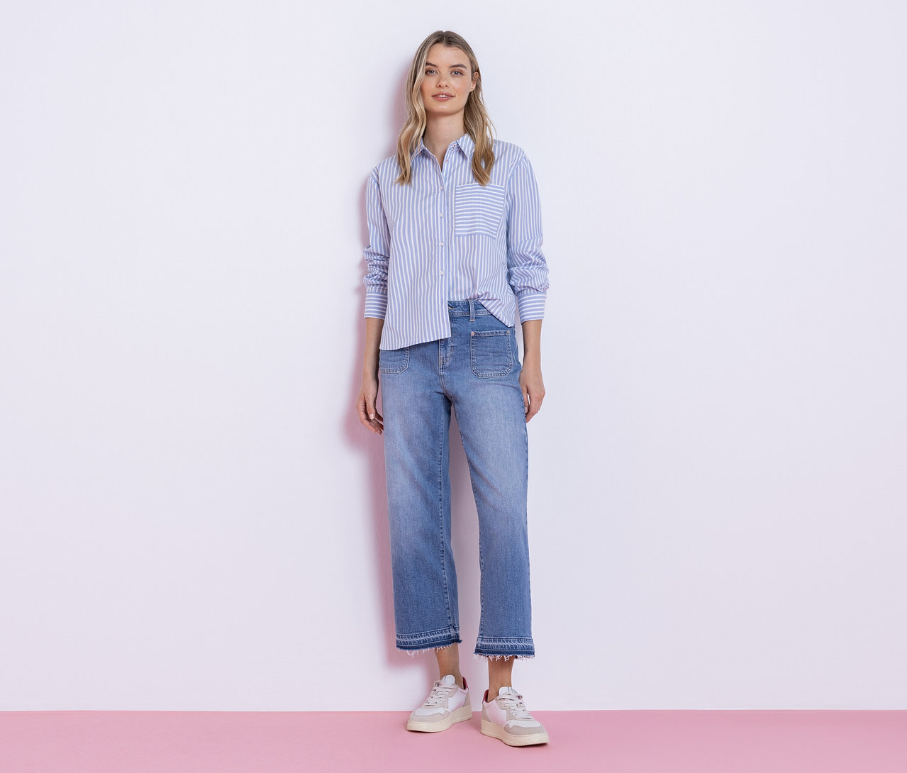 Młoda kobieta stoi przed białą ścianą, ubrana w niebiesko-białą koszulę Street One w paski, jasne dżinsy z postrzępionymi nogawkami i białe sneakersy.