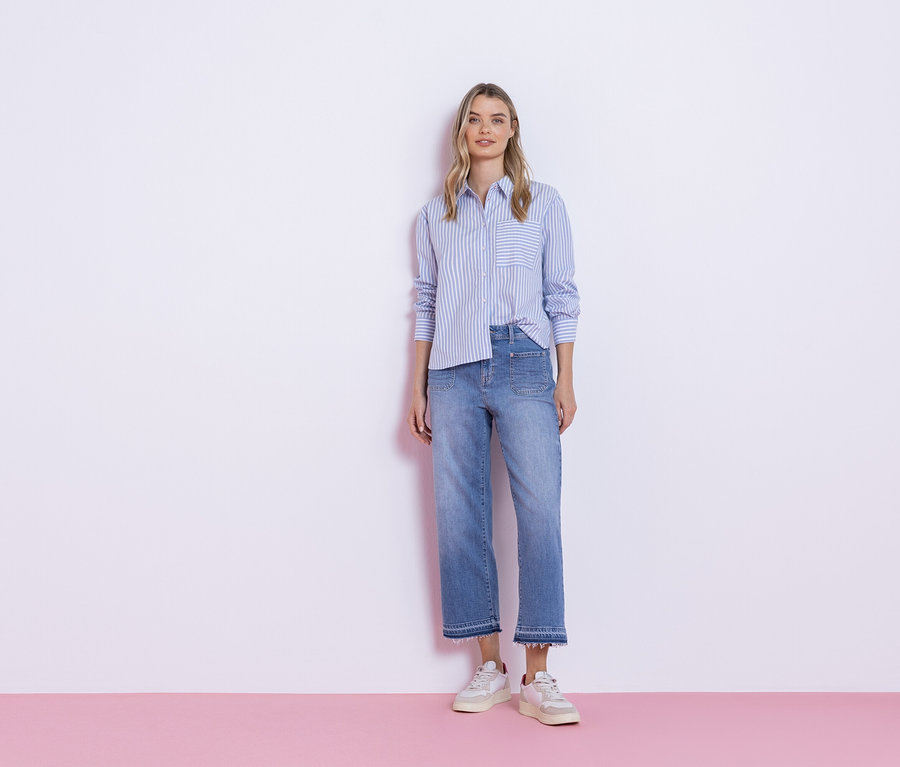Kobieta stoi na tle białej ściany, ubrana w niebieską koszulę w paski, dżinsy i trampki.