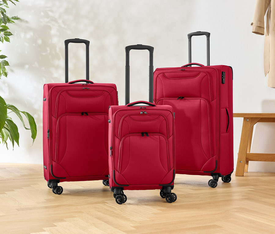 Trzy czerwone walizki na kółkach o różnych rozmiarach stoją na drewnianej podłodze, obok rośliny i ławki.