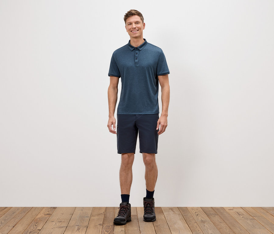 Mężczyzna stoi na tle białej ściany, ubrany w niebieską koszulkę polo, krótkie spodenki i buty trekkingowe.