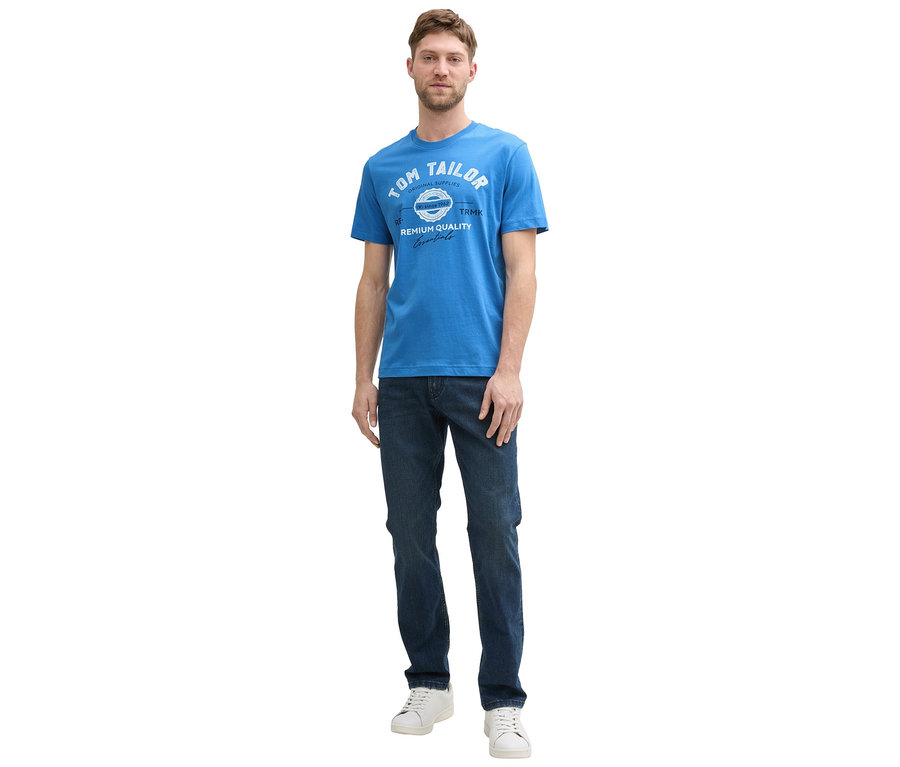 Mężczyzna w dżinsach i białych butach sportowych ma na sobie koszulkę z logo Tom Tailor w kolorze sapphire blue.