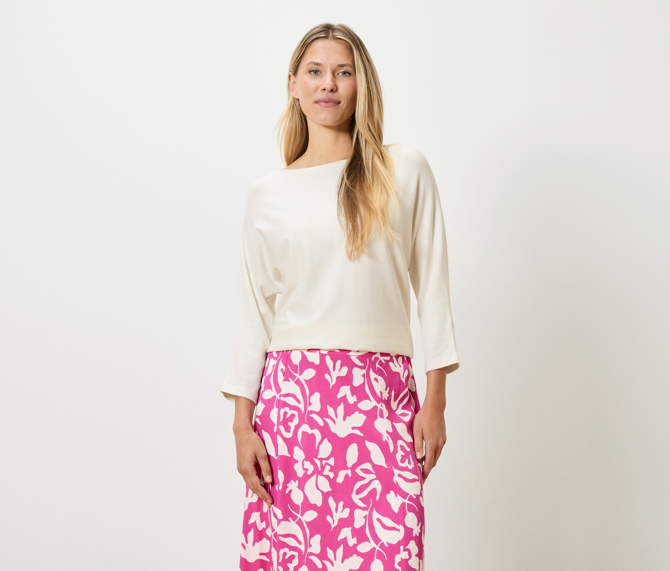 Kobieta o blond włosach ma na sobie kremową bluzkę i różową spódnicę w kwiaty.
