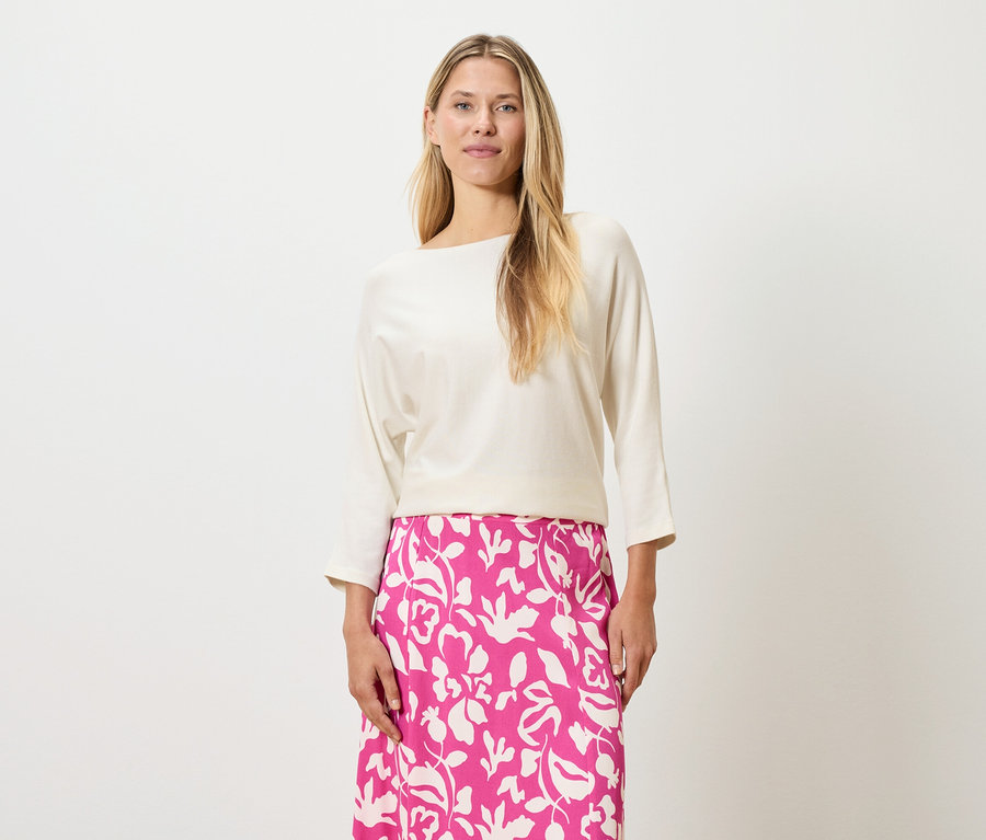 Kobieta o blond włosach ma na sobie kremową bluzkę i różową spódnicę w kwiaty.