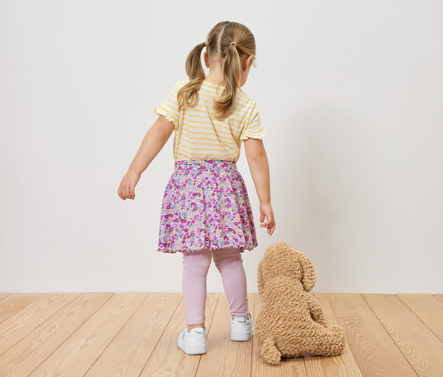 Widok z tyłu na dziewczynkę w warkoczach, ubraną w bluzkę w żółto-białe paski, kwiecistą spódnicę, legginsy i tenisówki, stojącą obok pluszowego misia.