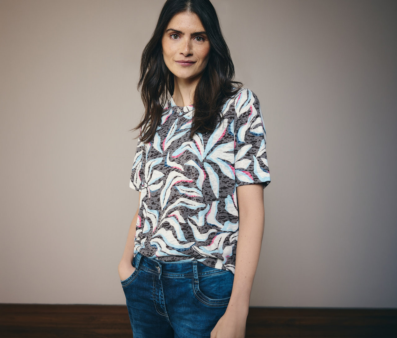 Kobieta o ciemnych włosach ma na sobie niebieską koszulkę z krótkim rękawem CECIL we wzory i niebieskie dżinsy.