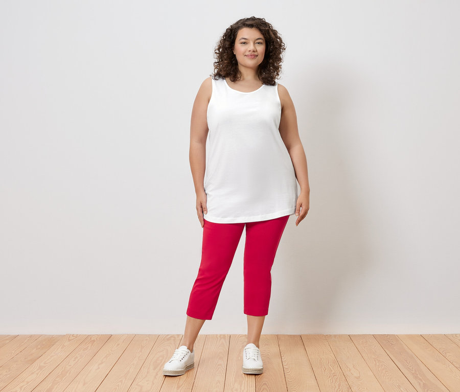 Kobieta z kręconymi włosami stoi na tle ściany, ubrana w biały top, czerwone spodnie i białe tenisówki.