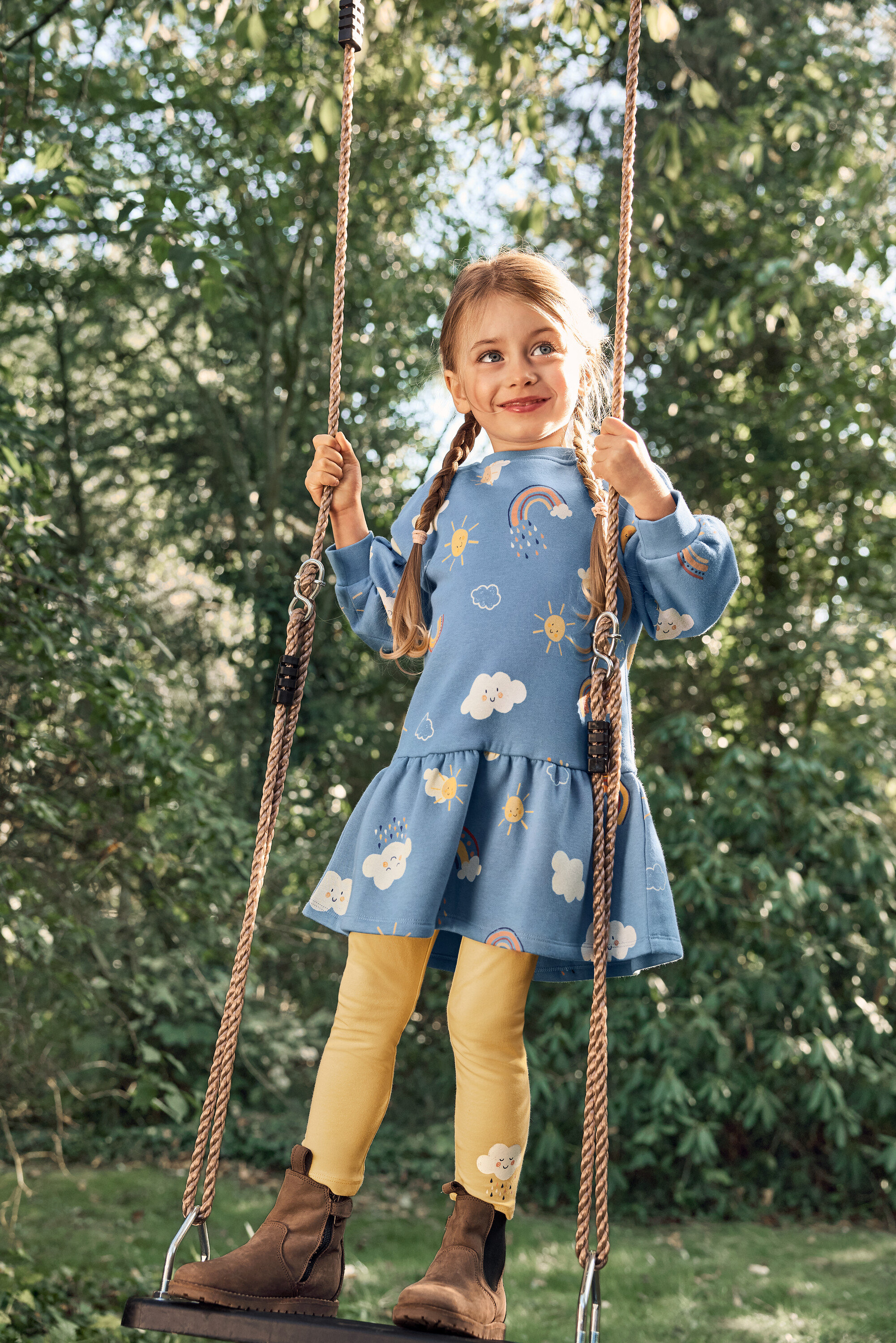 Dziewczynka w warkoczach huśta się na huśtawce, ubrana w niebieską sukienkę we wzory słońca, chmur i tęczy, żółte legginsy i brązowe buty.