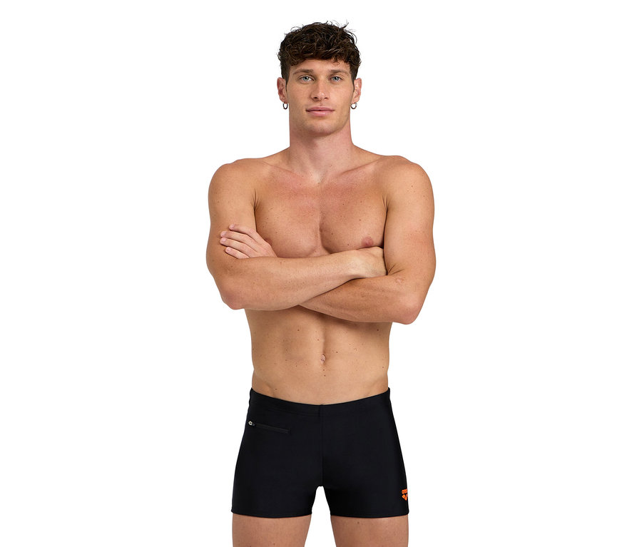 Mężczyzna ze skrzyżowanymi ramionami, ubrany w czarne kąpielówki.