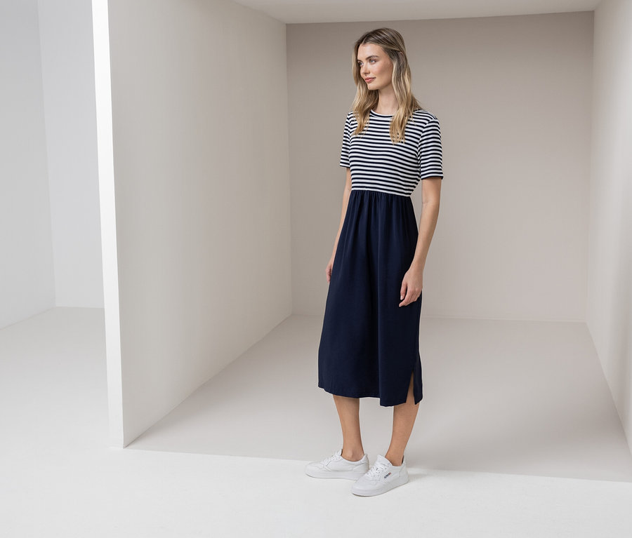 Kobieta stoi w białym pokoju, ubrana w sukienkę w paski biało-niebieskie i białe tenisówki.