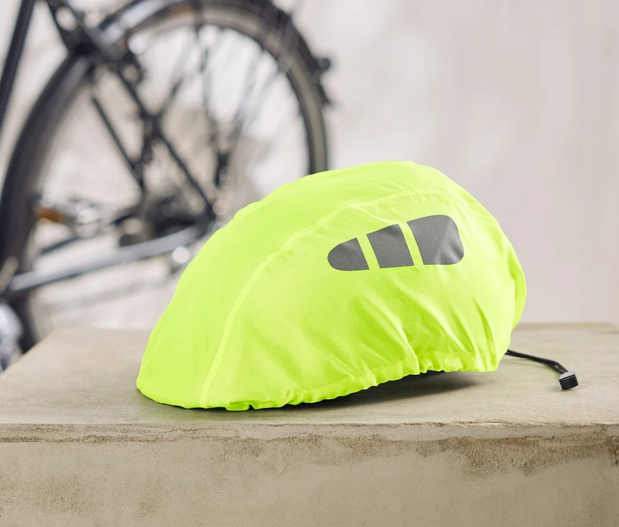 Neonowożółty pokrowiec na kask rowerowy z odblaskowymi paskami leży na betonowym cokole, w tle rower.