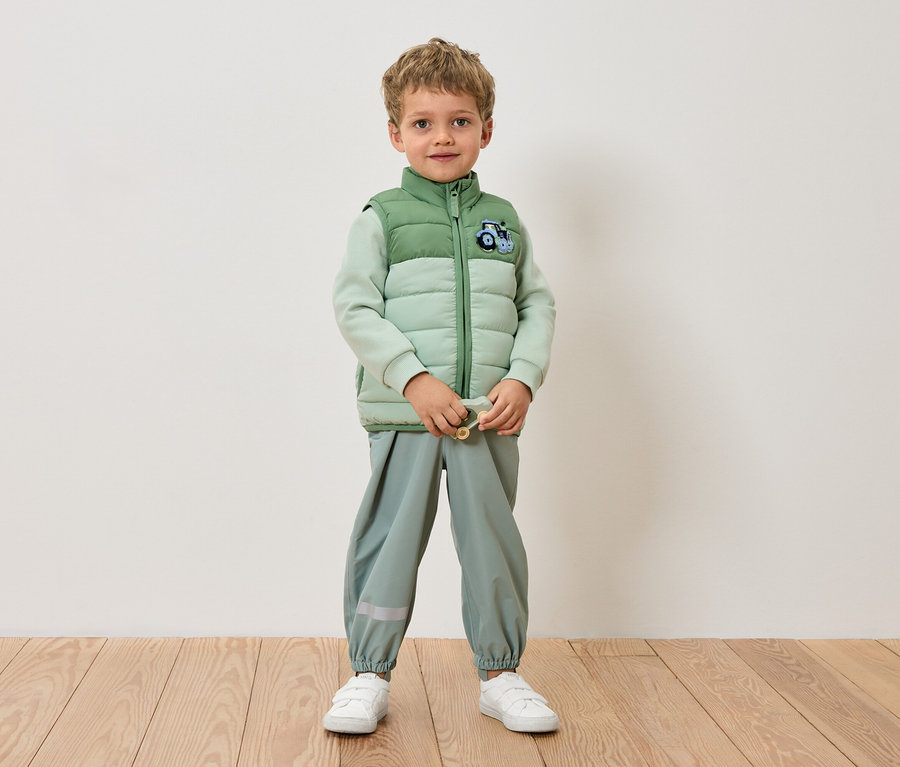 Chłopiec stoi na tle ściany w jasnozielonej kurtce, jasnozielonych spodniach i białych tenisówkach.