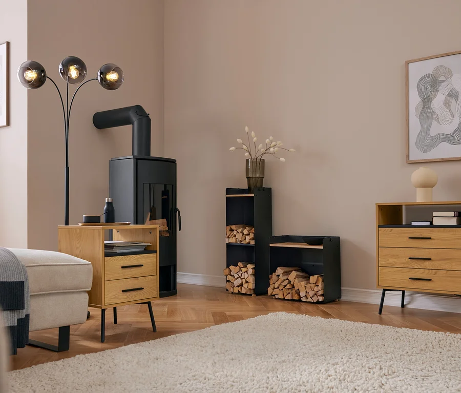 Nowoczesny salon z czarnym piecem kominkowym, wysoką szafką na drewno do kominka z blatem i szeroką szafką na drewno do kominka z blatem, obie wypełnione drewnem opałowym. Jasna sofa, drewniany stolik i komoda stoją na dywanie.