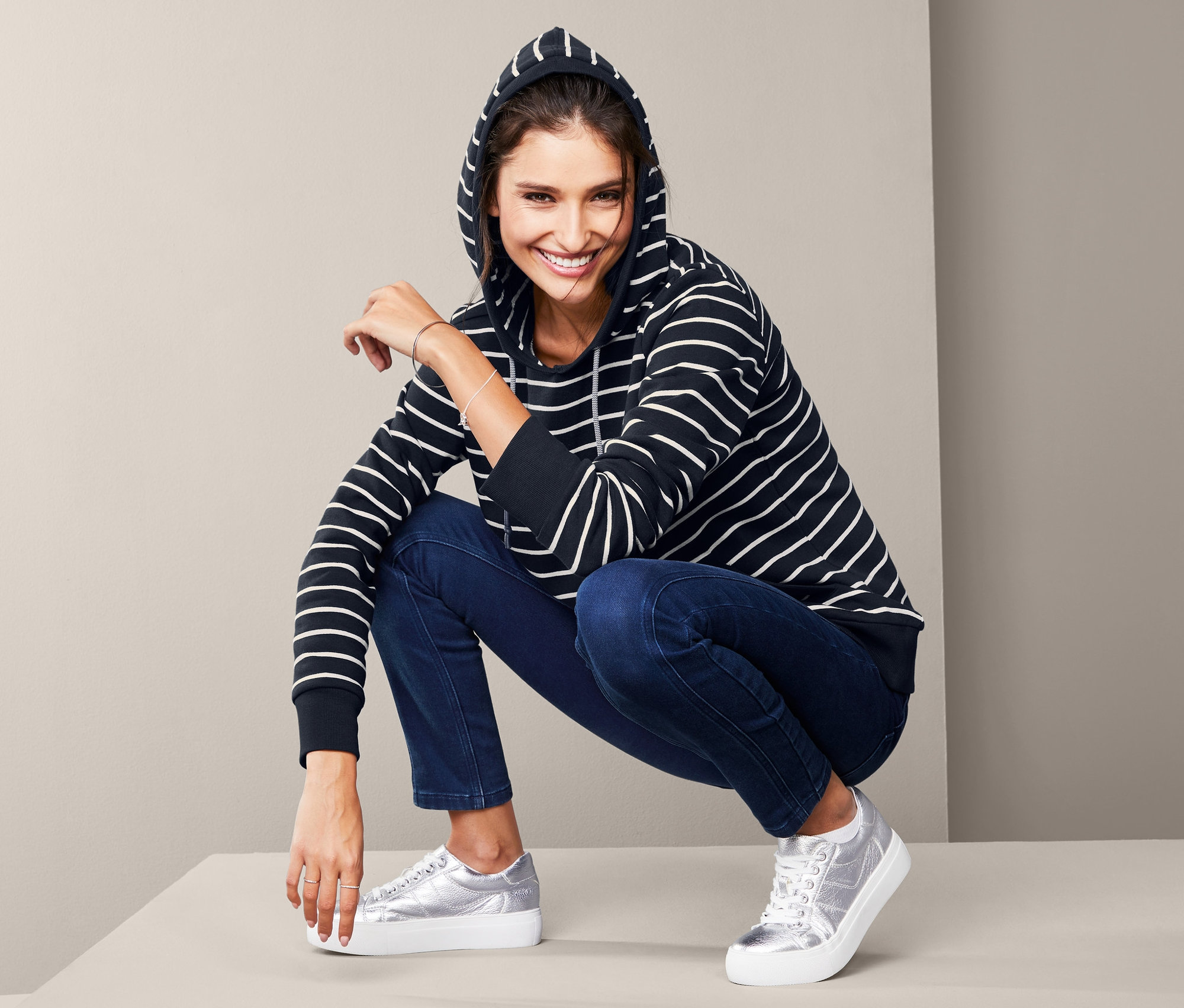 Kobieta w bluzie damskiej z kapturem w paski i dżinsach pozuje na podłodze.