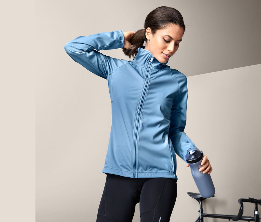 Kobieta w kurtce softshell na rower trzyma butelkę z wodą obok roweru.