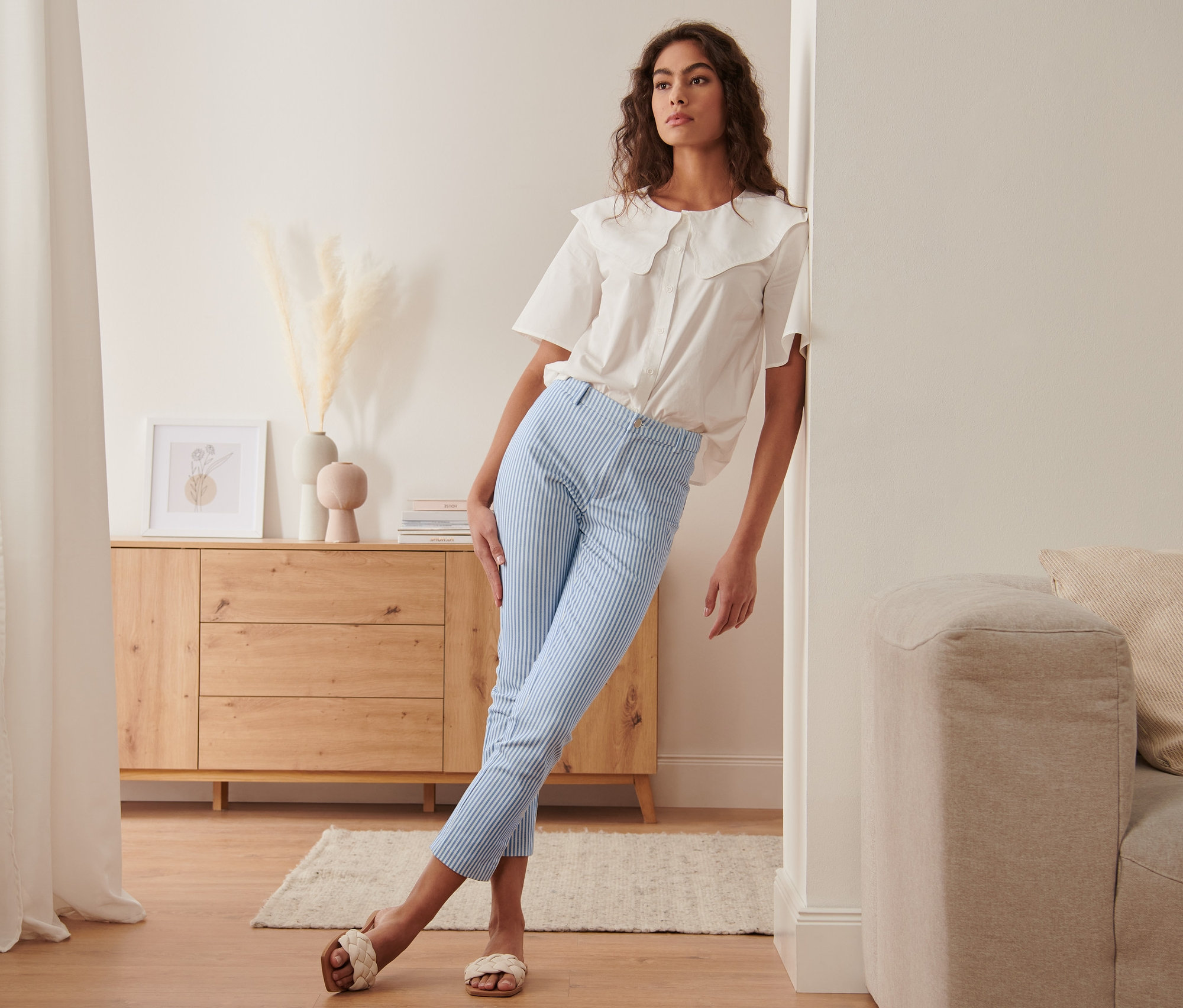 Kobieta opiera się o ścianę, ma na sobie pasiaste spodnie ze stretchu i białą bluzkę.