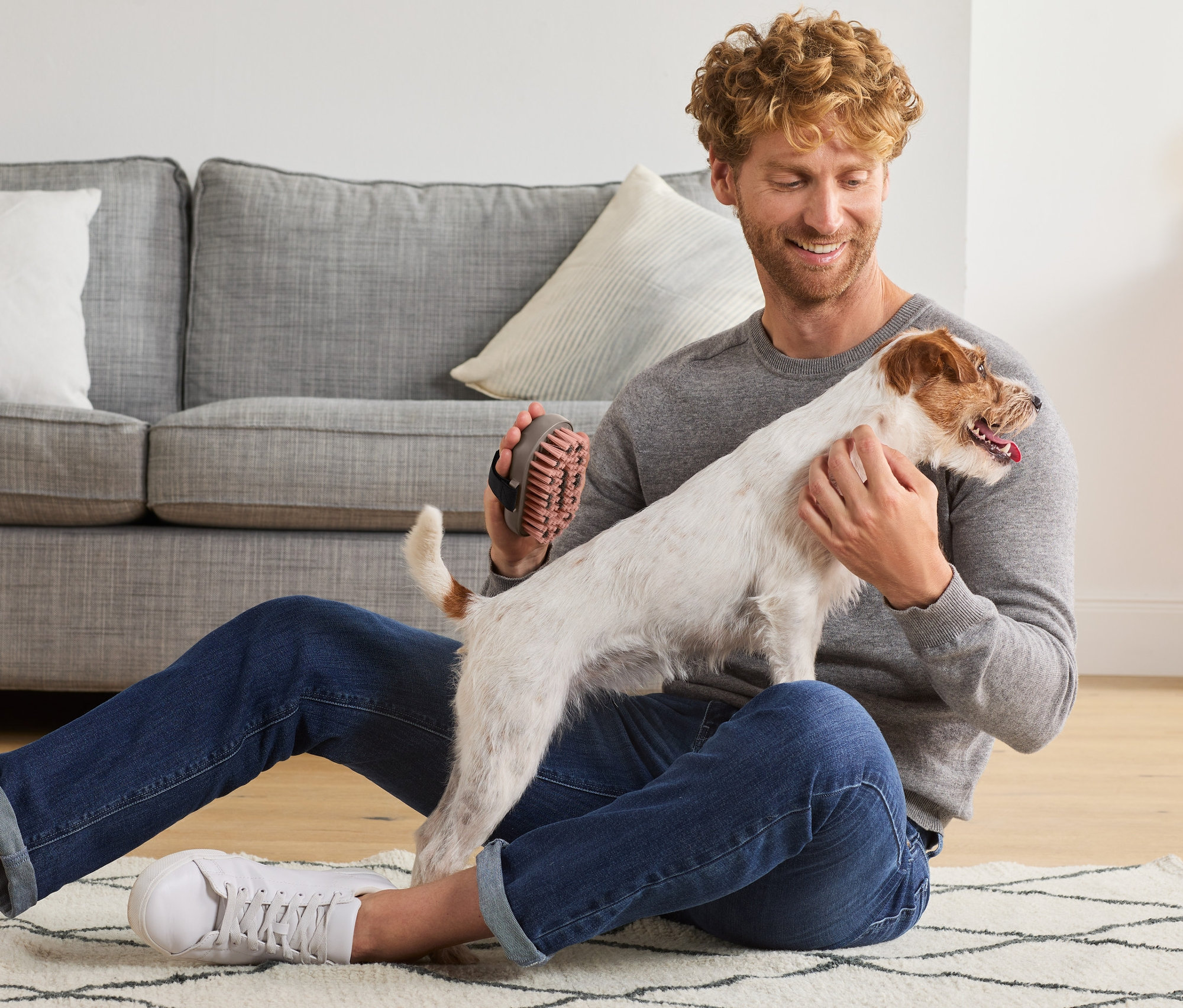 Mężczyzna siedzi na podłodze i trzyma psa, w ręku ma szczotkę do krótkiej sierści.