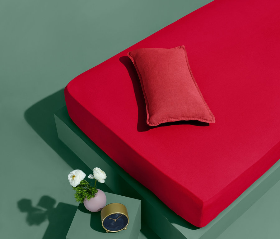 Czerwone prześcieradło dżersejowe z gumką w standardowym rozmiarze i czerwona poduszka na zielonym tle.