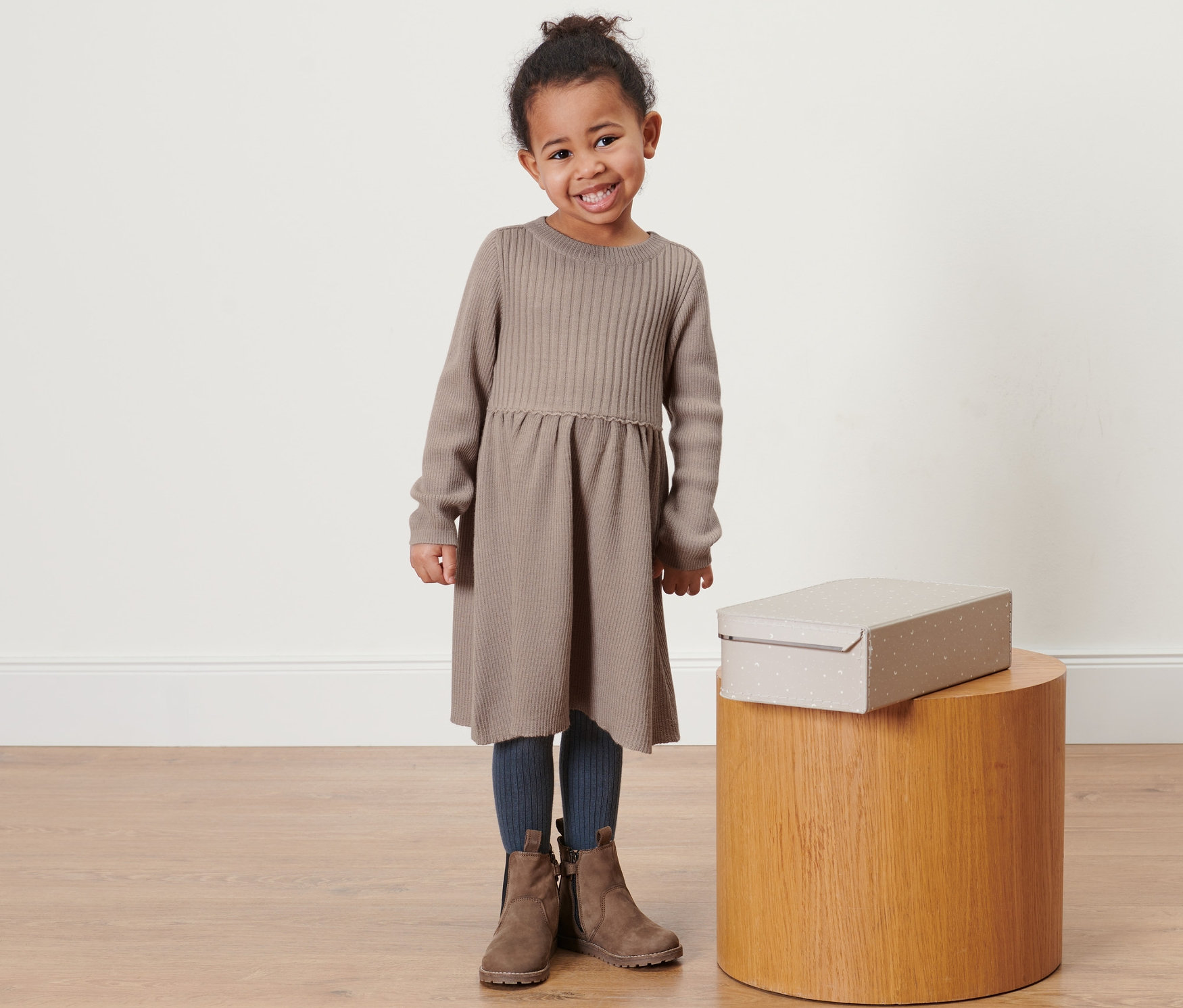 Uśmiechnięta dziewczynka pozuje w sukience z dzianiny z zawartością wełny merino w kolorze jasnobrązowym. Obok niej na drewnianym stołku stoi pudełko.