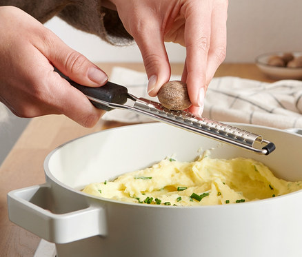 Kobieta trze gałkę muszkatołową na tarce do gałki muszkatołowej nad garnkiem z puree.