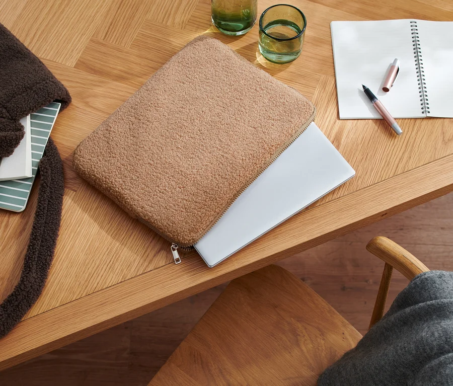Na drewnianym stole leżą pluszowa torba na laptopa, notatnik, otwarty notes, długopis i szklanki.