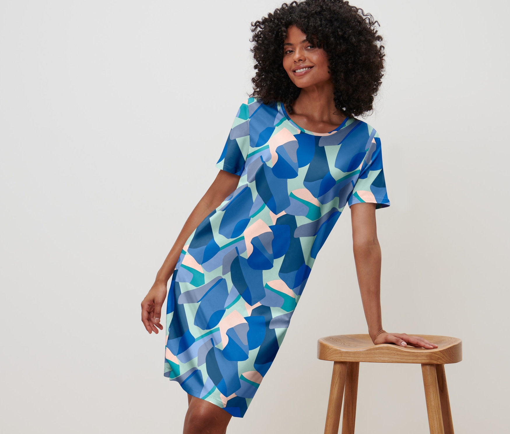 Kobieta pozuje w niebieskiej koszuli nocnej we wzory, opierając się o drewniany stołek.