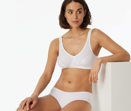 Kobieta siedzi i ma na sobie białą braletkę z miseczkami i bawełną ekologiczną SCHIESSER, 2 sztuki.