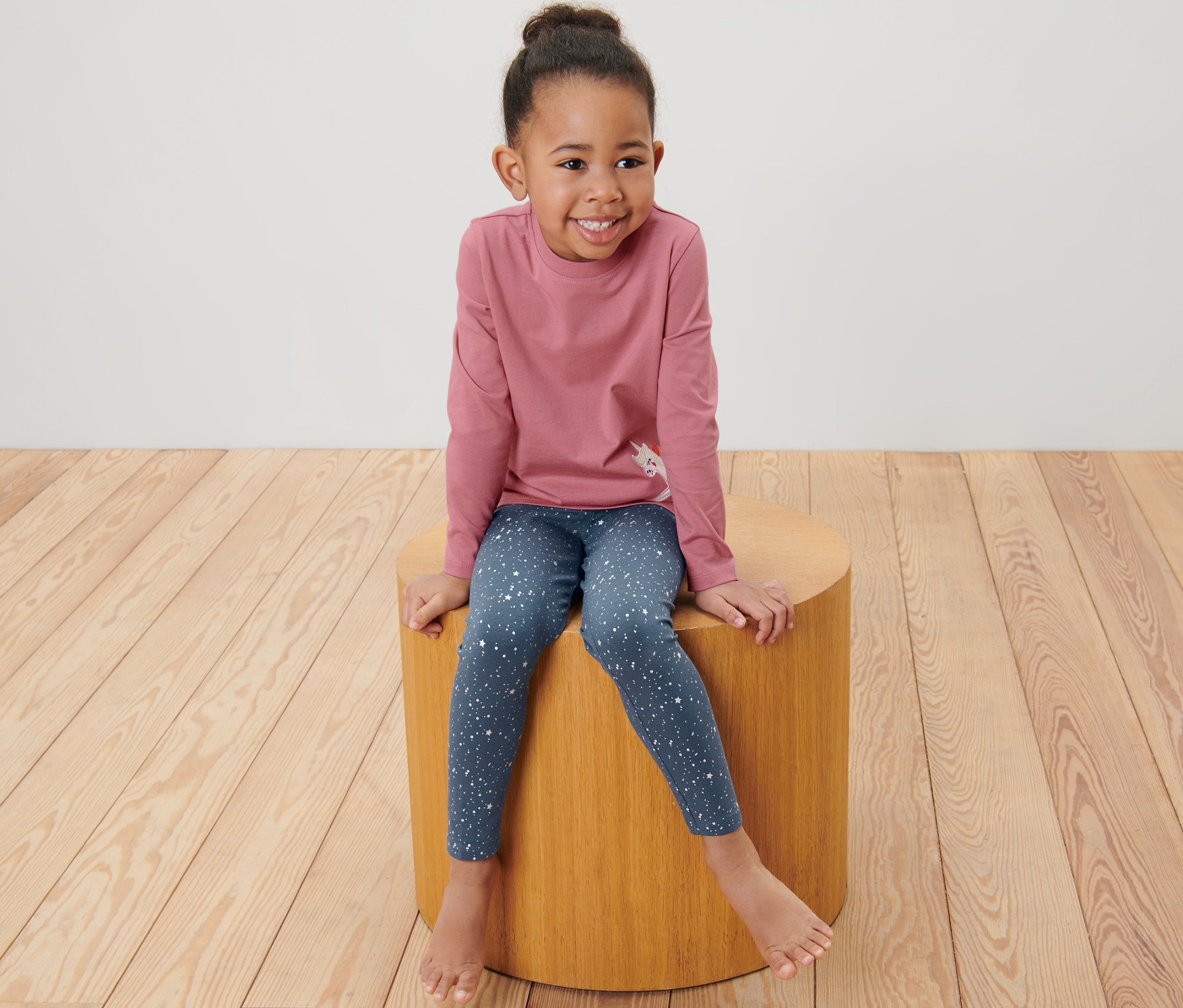 Uśmiechnięta dziewczynka siedzi na drewnianym stołku i ma na sobie dziecięce legginsy z brokatem, 2 pary.