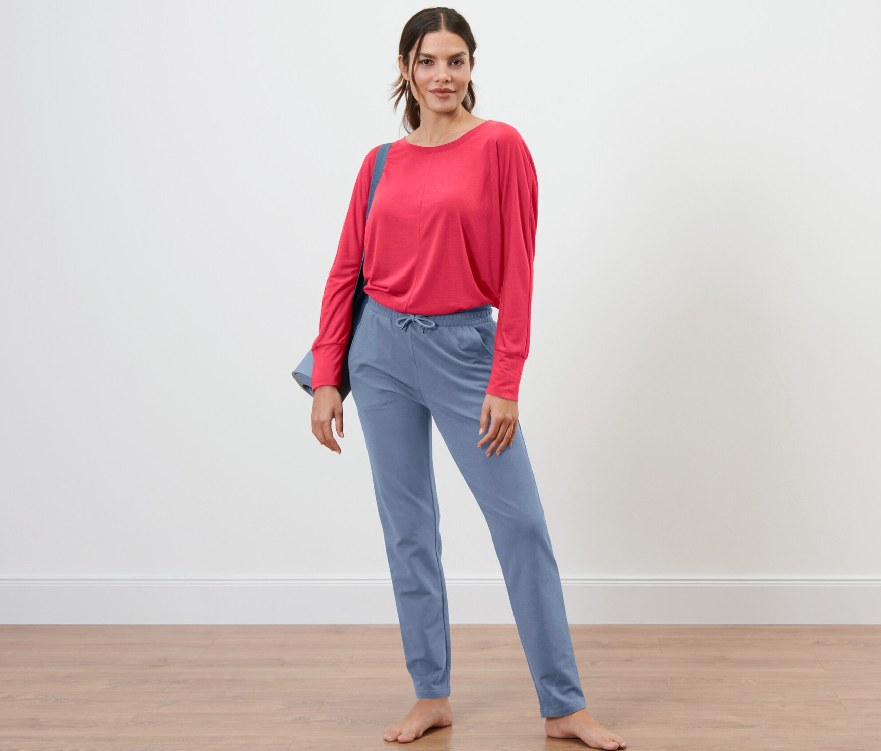 Kobieta stoi boso, ubrana w szaroniebieskie spodnie dresowe i czerwoną bluzkę.
