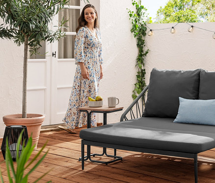 Kobieta w sukience stoi obok stolika z aluminium w kształcie litery C i rozsuwanej sofy wypoczynkowej. W tle widoczny solarny łańcuch świetlny, rowkowane kule.