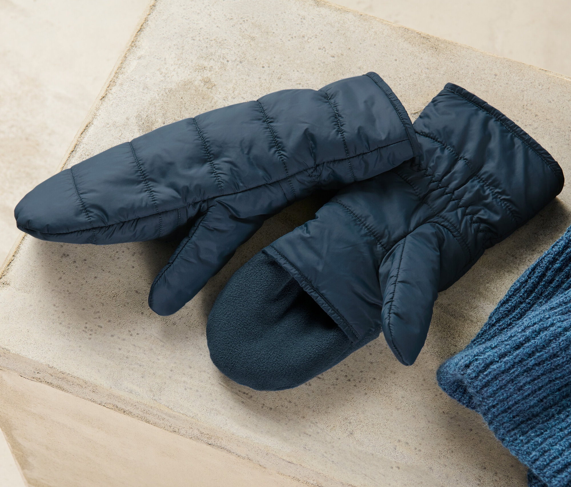 Dwie granatowe pikowane rękawice jednopalczaste z klapką leżą na szarej powierzchni obok niebieskiej czapki z dzianiny.