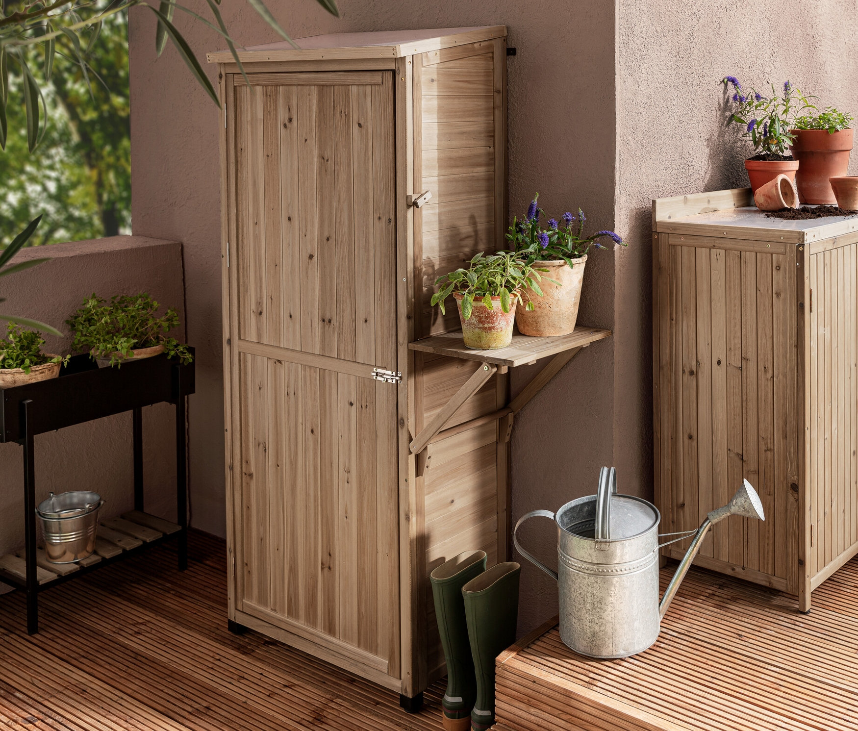Dwie szafy ogrodowe „Tinus”, jedna duża ze składaną półką boczną i jedna mała, oraz stojak na rośliny „Elin” na drewnianym tarasie.