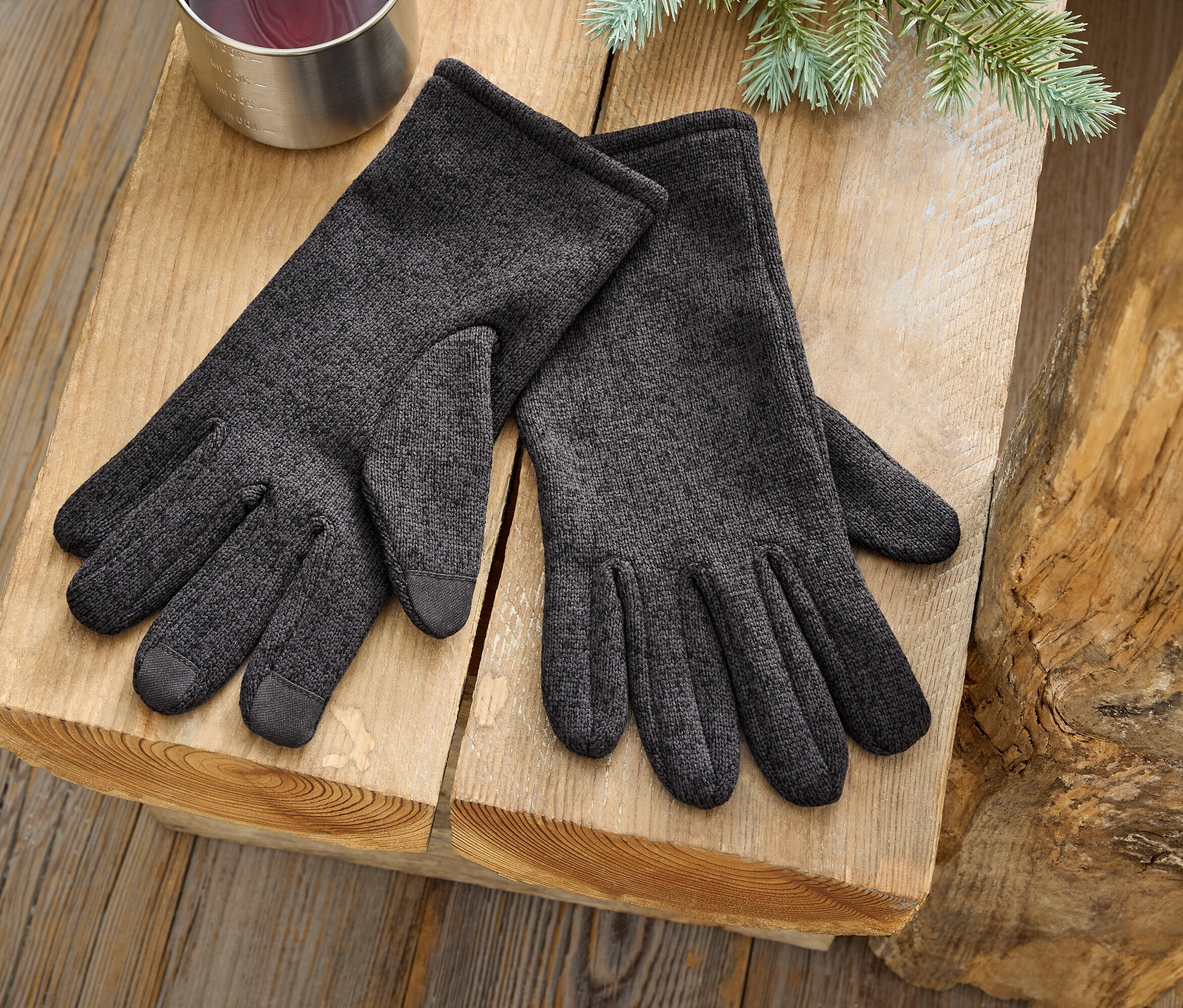 Para szarych rękawic leży na drewnianej powierzchni obok srebrnego kubka i gałązki drzewa iglastego.