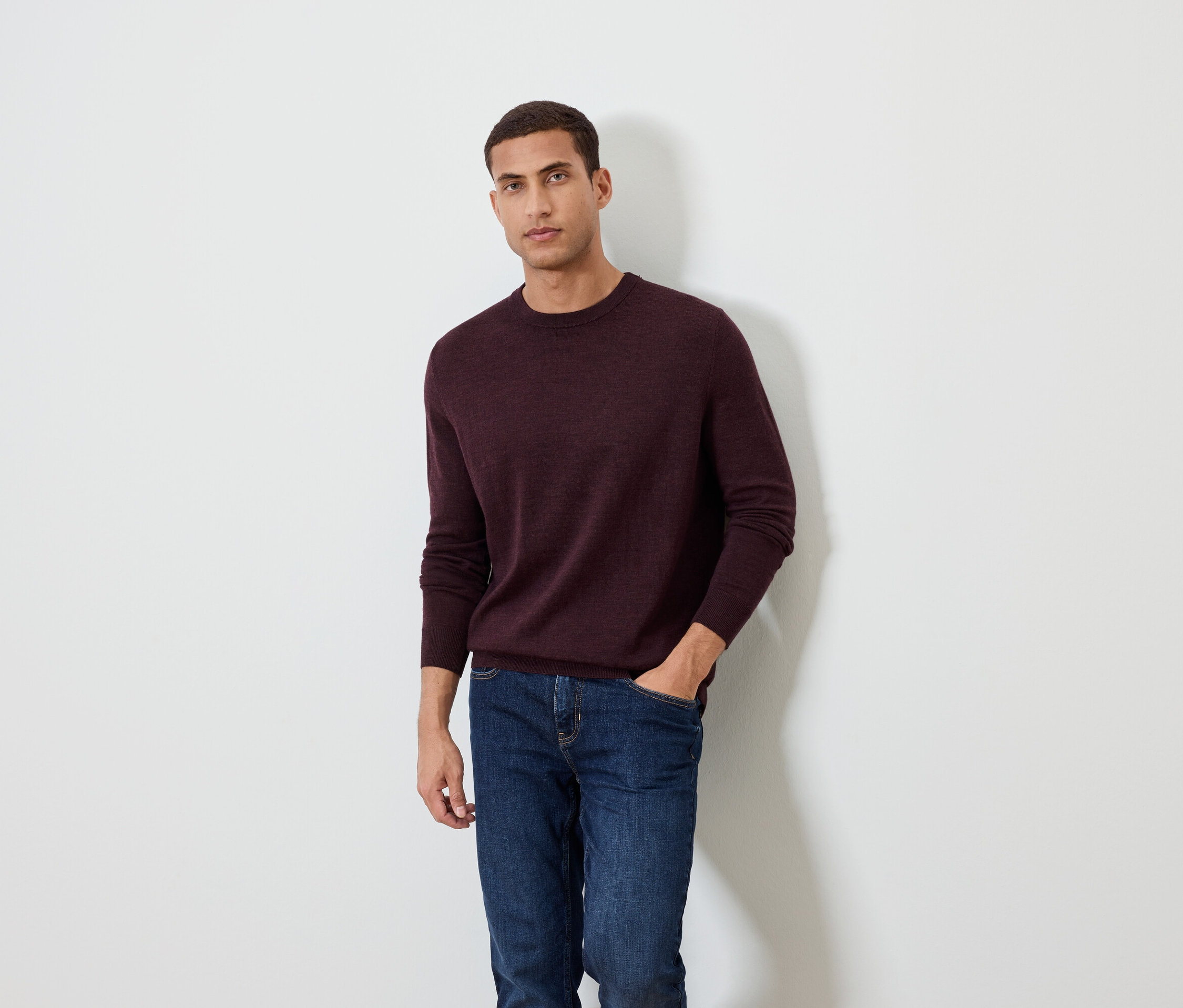 Mężczyzna pozuje na białym tle, ubrany w bordowy sweter i dżinsy.