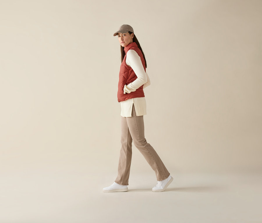 Kobieta idzie, ubrana w kamizelkę pikowaną, bluzkę z długim rękawem, spodnie i czapkę z daszkiem.