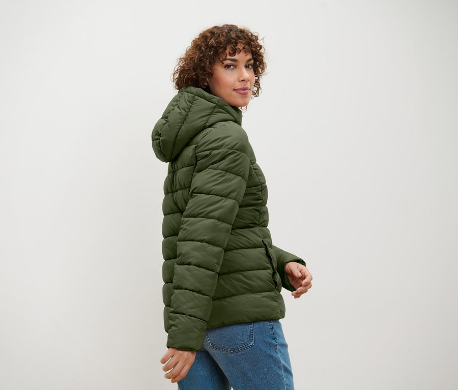 Kobieta z kręconymi włosami spogląda przez ramię, pozując w oliwkowozielonej kurtce puchowej z kapturem i kurtce jeansowej.