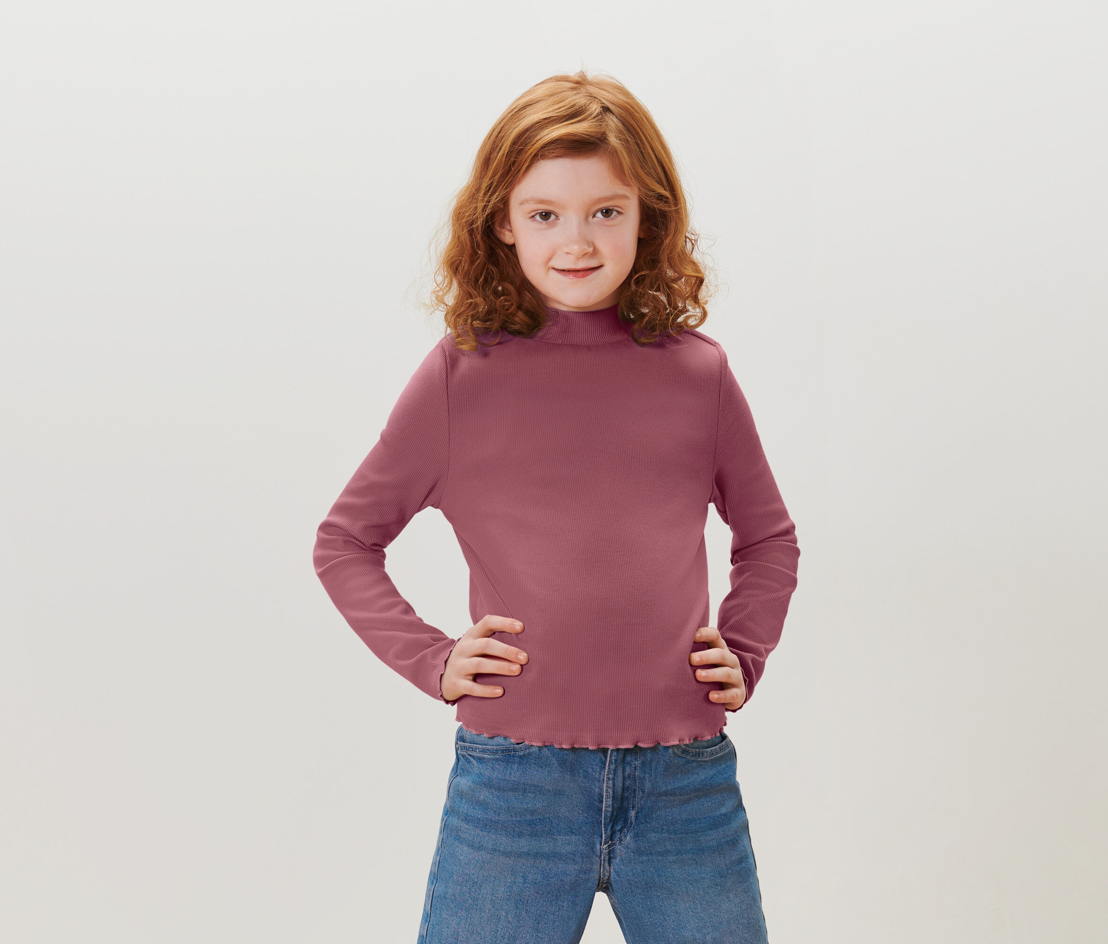Dziewczyna z rudymi kręconymi włosami stoi z rękami na biodrach. Ma na sobie purpurową koszulkę z długim rękawem i niebieskie dżinsy.