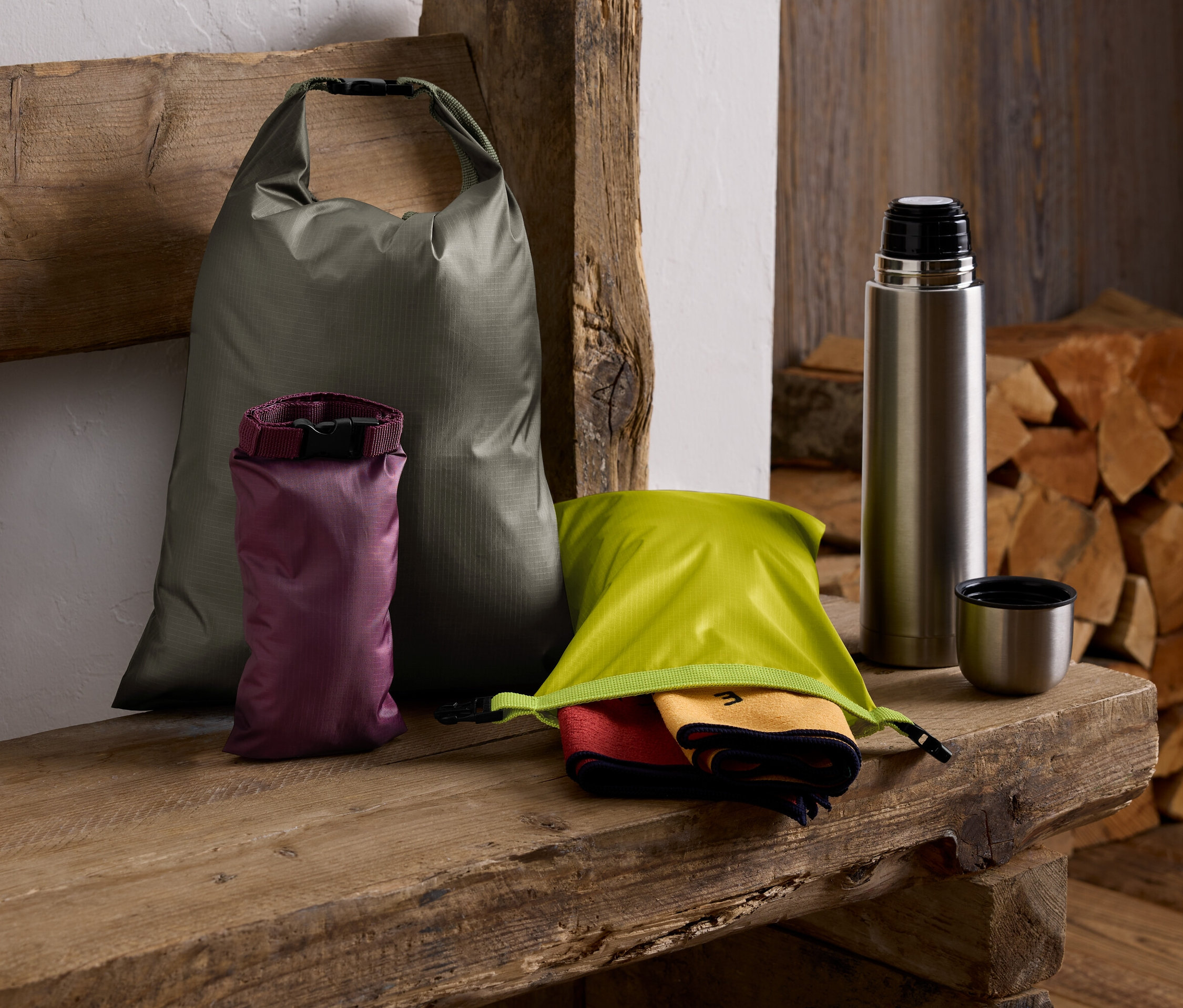 Na drewnianej ławce leżą zielony worek, fioletowy worek, zielony worek z ręcznikami, termos i stos drewna.