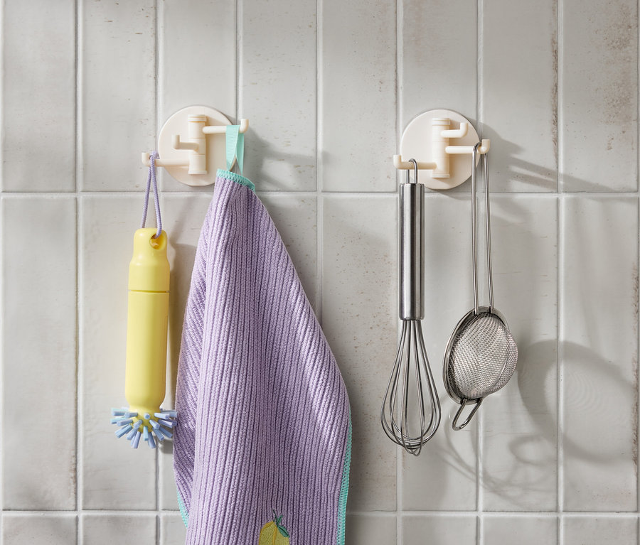 Na białych kafelkach wiszą żółta szczotka, fioletowa ścierka, trzepaczka ze stali nierdzewnej i sitko.