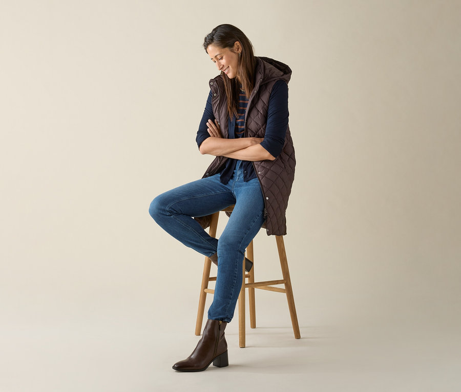 Kobieta siedzi na stołku ze skrzyżowanymi ramionami i ma na sobie długą pikowaną kamizelkę z kapturem w kolorze ciemnobrązowym.