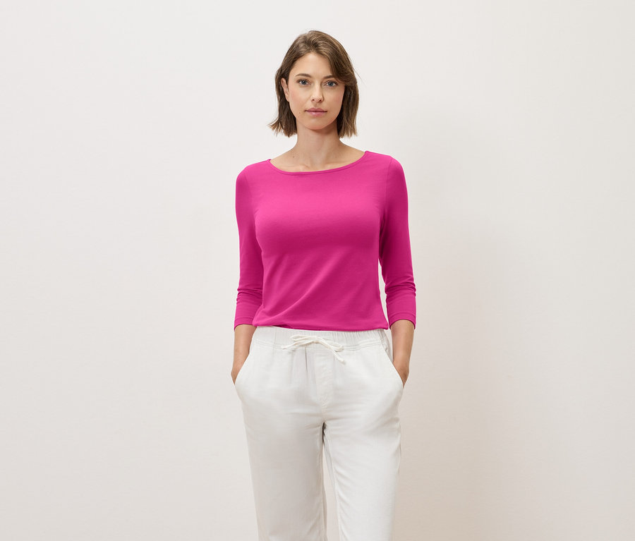 Kobieta z brązowymi włosami pozuje w różowym topie z rękawami trzy czwarte i białych spodniach.