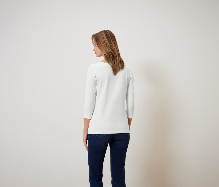 Kobieta z brązowymi włosami stoi tyłem, ubrana w białą koszulę z rękawami 3/4 i niebieskie dżinsy.