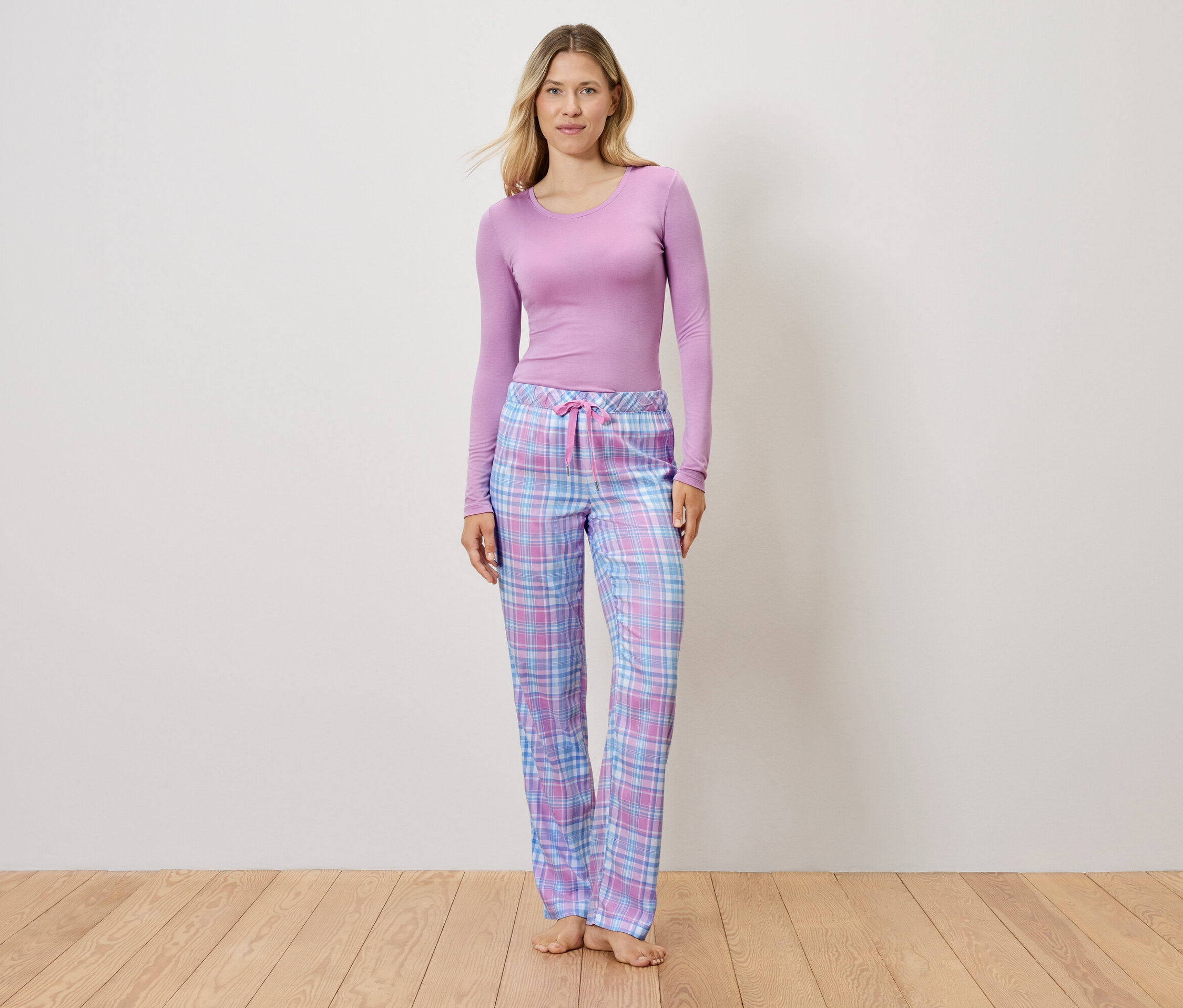 Kobieta w liliowym topie i spodniach od piżamy w kratę stoi na drewnianej podłodze.