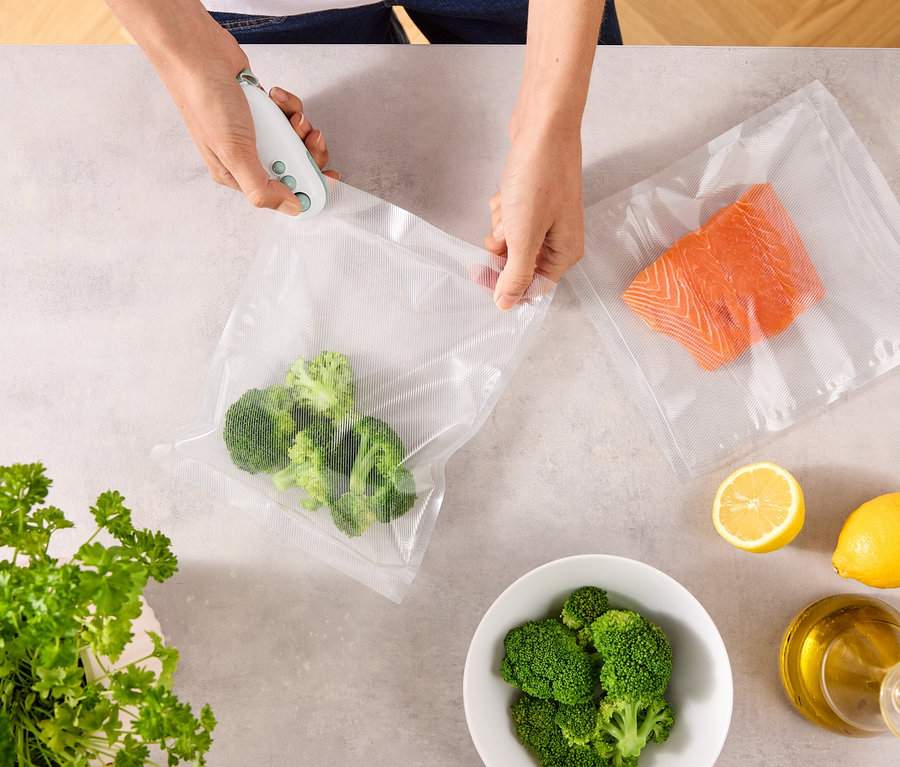 Osoba zamyka worek z brokułami za pomocą zgrzewarki próżniowej. Na stole leżą: drugi worek z łososiem, cytryny, miska brokułów, pietruszka i butelka oleju.