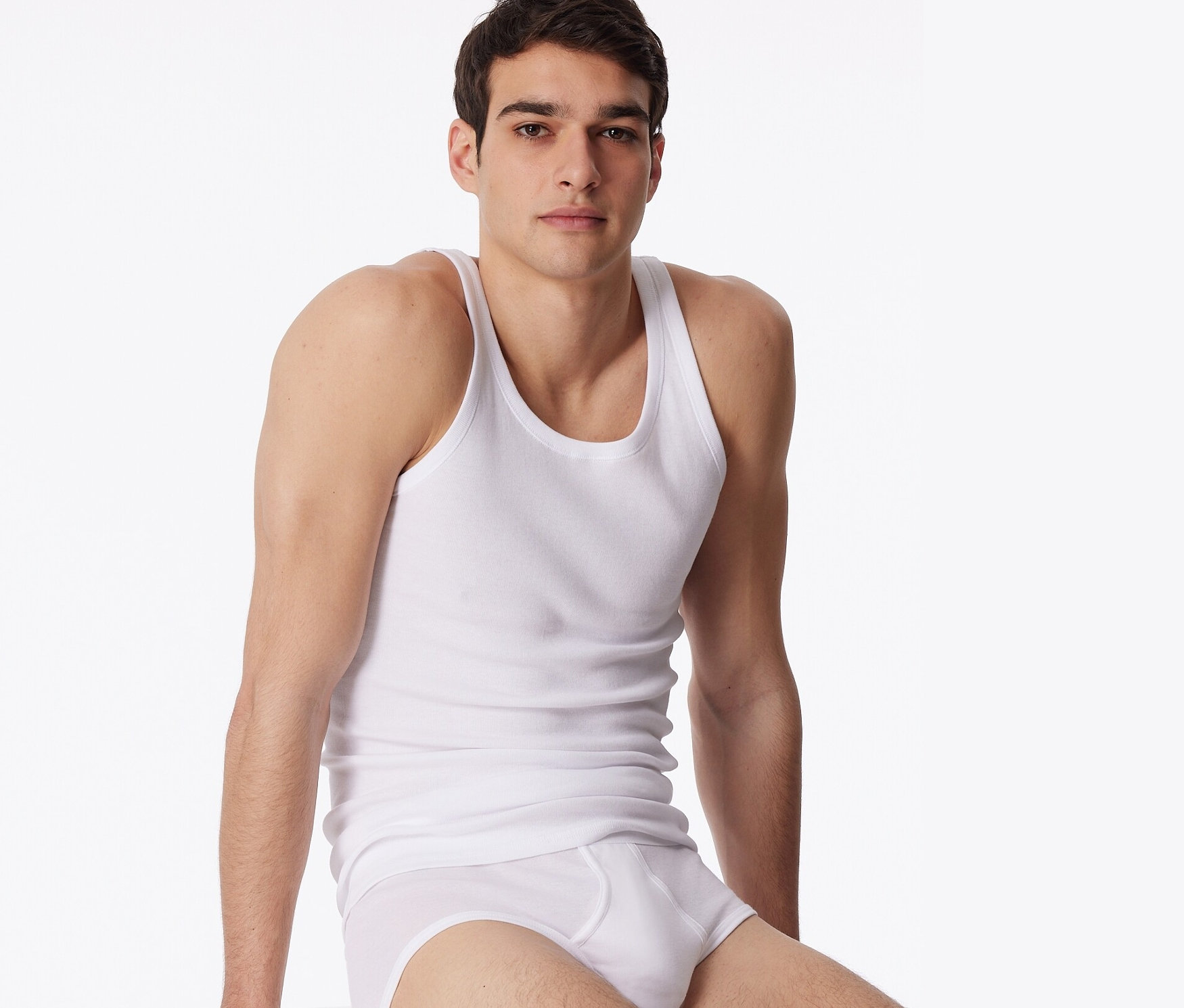 Mężczyzna siedzi na białym sześcianie, ubrany w białą bieliznę i białą podkoszulkę trykotową SCHIESSER, 2 sztuki.