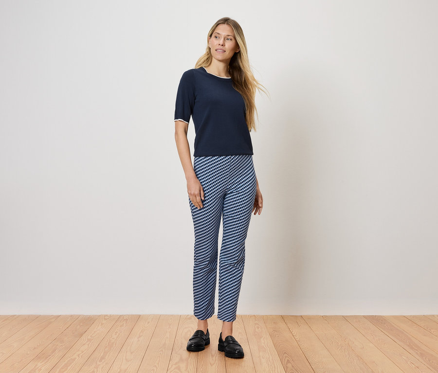 Kobieta stoi na drewnianej podłodze, ubrana w granatową koszulkę i niebiesko-białe spodnie w paski.